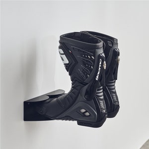 Puede incluir: Un par de botas de moto negras con detalles blancos y la palabra "Dainese" impresa en el lateral. Las botas están montadas en un soporte de botas de pared negro.