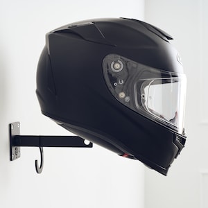 Può includere: Un casco da moto nero con una visiera trasparente è appeso a un gancio di metallo nero montato su una parete bianca.