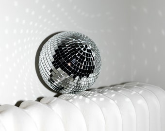 May include: A silver disco ball with mirrored tiles sits atop a white radiator. The ball reflects light onto the white wall, creating a pattern of small, square light spots. The image evokes a party or celebration theme.