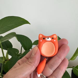 May include: A handmade orange fox charm with a white belly and tail tip. The fox charm has a small black eye and a light orange belly. The fox tail is attached to the fox charm with a small silver ring. The charm is held in a hand.