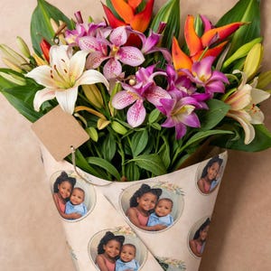 May include: A vibrant bouquet of lilies, orchids, and other colorful flowers. The bouquet is wrapped in paper decorated with portraits of a smiling girl and a baby. A blank brown tag hangs from the bouquet.
