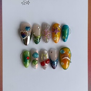 May include: A collection of ten artificial nails with diverse designs. The nails are adorned with embellishments such as hearts, glitter, and pearls. The card displays the text "alanailz" and "handmade in france".