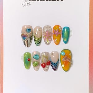 May include: A set of ten artificial nails with diverse designs, including hearts, glitter, floral accents, and a star. The nails are in various colours, such as green, orange, and blue. The text "alanailz" and "handmade in france" are visible.