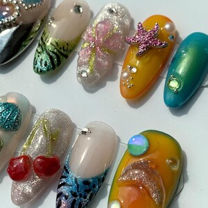 May include: A selection of ten long, decorated artificial nails. Designs include glitter, rhinestones, and various shapes, such as a starfish, cherries, and a dolphin. Colours include pink, yellow, blue, and green.