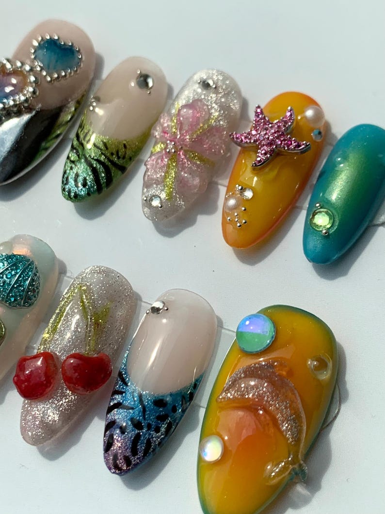 May include: A selection of ten long, decorated artificial nails. Designs include glitter, rhinestones, and various shapes, such as a starfish, cherries, and a dolphin. Colours include pink, yellow, blue, and green.