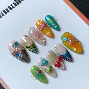 May include: A selection of artificial nails with diverse designs. The nails showcase a variety of colours, including green, yellow, and silver, and are decorated with embellishments like glitter, pearls, and starfish. The nails are presented on a white surface.