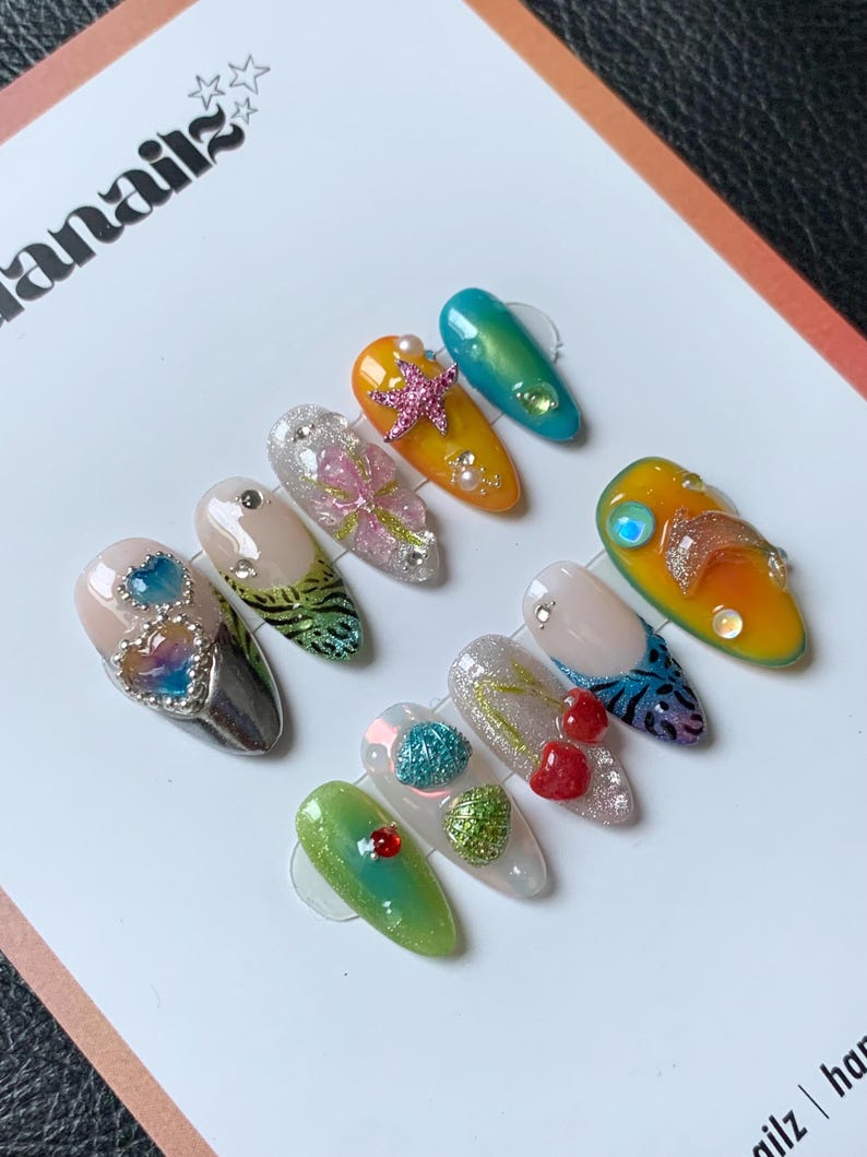 May include: A selection of artificial nails with diverse designs. The nails showcase a variety of colours, including green, yellow, and silver, and are decorated with embellishments like glitter, pearls, and starfish. The nails are presented on a white surface.