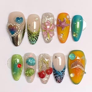 May include: A collection of ten artificial nails with diverse designs. The nails showcase a variety of colours, including silver, pink, green, and orange. Some nails are adorned with embellishments such as hearts, flowers, and cherries. The nails are presented on a white surface.