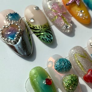 May include: A selection of artificial nails with diverse designs. Some nails feature heart-shaped gems, while others showcase floral patterns, starfish, and other decorative elements. The nails are in various colours, including pink, green, and orange.