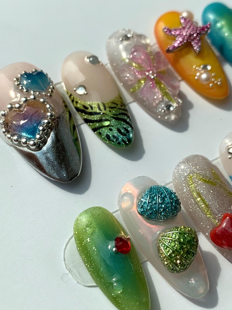 May include: A selection of artificial nails with diverse designs. Some nails feature heart-shaped gems, while others showcase floral patterns, starfish, and other decorative elements. The nails are in various colours, including pink, green, and orange.