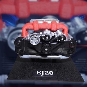 May include: A detailed model of an engine, featuring red intake manifolds and black components. The model sits on a black base with the text "EJ20". The engine's silver and black parts are visible, showcasing belts and pulleys.