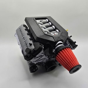 May include: A 3D printed model of a Ford V8 engine featuring a black engine cover with the text 5.0, a silver valve cover with the text POWERED BY FORD, and a prominent red conical air filter attached to the front intake assembly.