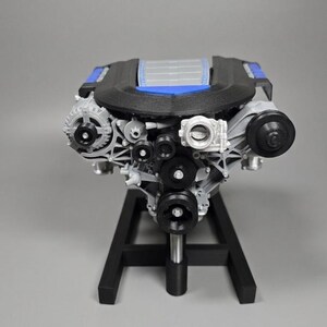 May include: A detailed model of an engine, featuring a black and blue top, gray components, and black pulleys. The engine is mounted on a black stand, showcasing its intricate design and mechanical elements.