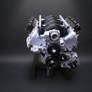 May include: A detailed model of a car engine, featuring a silver and black color scheme. The engine is mounted on a dark gray stand, showcasing its intricate components. The design includes multiple pulleys and belts.