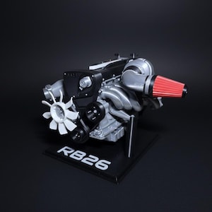 May include: A detailed model of an RB26 engine, featuring a silver and black color scheme with a red air filter. The model is mounted on a black base with the text "RB26" in white. The engine includes a fan and various mechanical components.