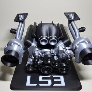 May include: A detailed model of an LS3 engine, featuring silver and black components. The model includes exhaust pipes, pulleys, and the text "LS3" in white on a black base. A realistic representation of an automotive engine.