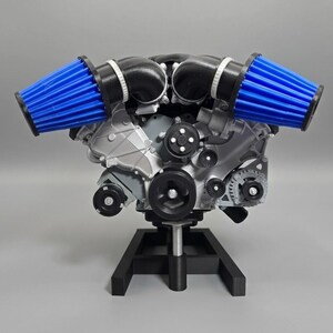May include: A detailed model of an engine with blue air filters. The engine is silver and black, mounted on a black stand. The blue filters are a striking contrast to the metallic components.