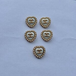 May include: Five heart-shaped buttons featuring a cream-colored center with a gold-toned interlocking CC logo. Each button is bordered by a gold-toned frame accented with a row of small white faux pearls arranged in a circular pattern around the edge.