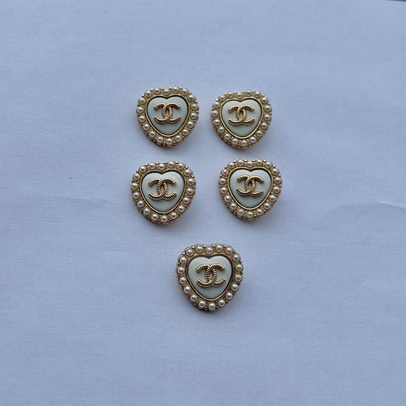 May include: Five heart-shaped buttons featuring a cream-colored center with a gold-toned interlocking CC logo. Each button is bordered by a gold-toned frame accented with a row of small white faux pearls arranged in a circular pattern around the edge.