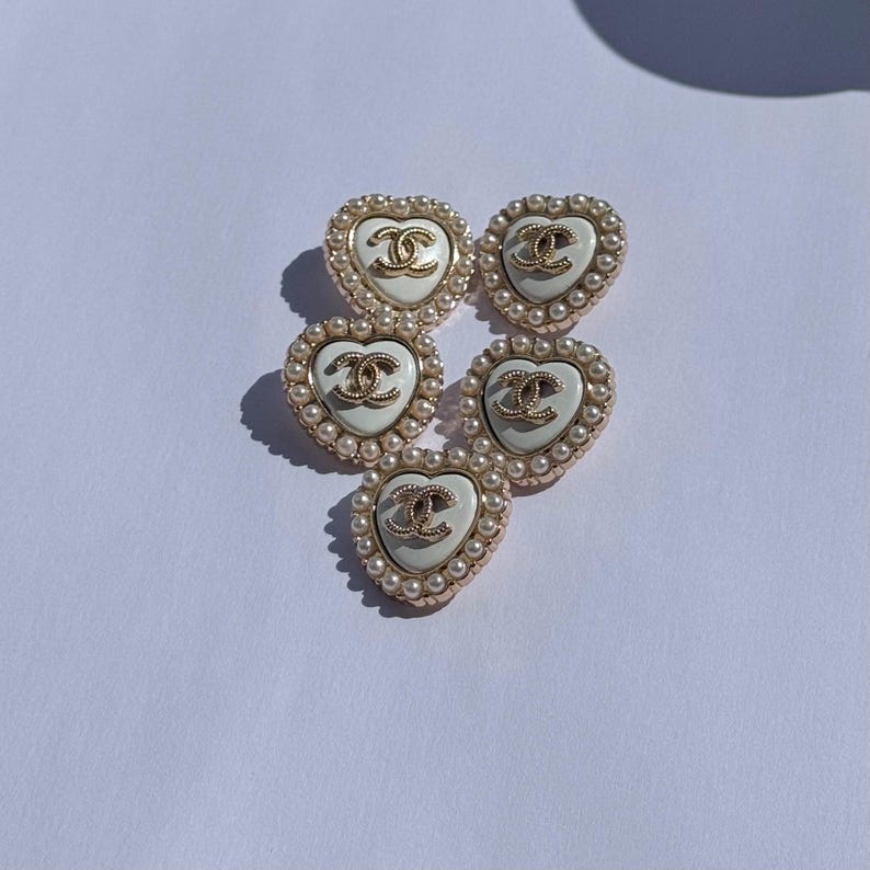 May include: Five heart-shaped buttons featuring a white center, a gold-toned interlocking CC logo, and a border of small white faux pearls. Each button has a metallic gold-toned edge surrounding the pearl trim, arranged in a cluster.