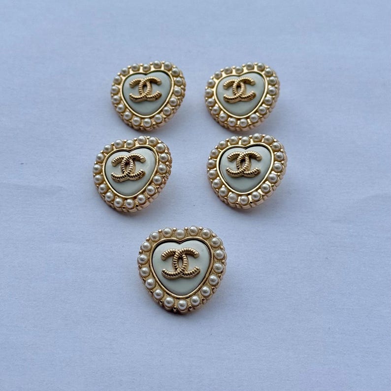 May include: Five heart-shaped buttons featuring a cream-colored center with a gold-toned interlocking CC logo. Each button is bordered by a gold-toned frame accented with small white faux pearls arranged in a circular pattern around the edge.