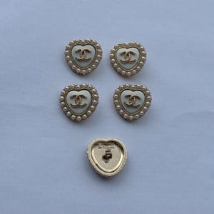 May include: Five heart-shaped buttons featuring a cream-colored center with a gold-tone interlocking CC logo. Each button is bordered by a row of small white faux pearls set in a gold-tone frame. One button is flipped to show the gold-tone back with CHANEL text.