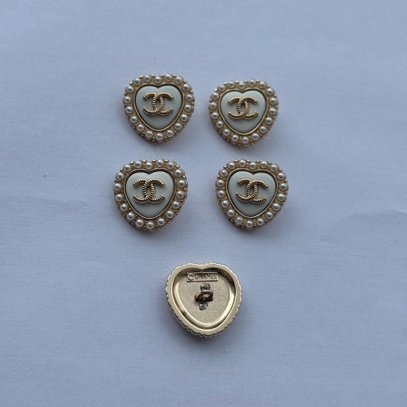 May include: Five heart-shaped buttons featuring a cream-colored center with a gold-tone interlocking CC logo. Each button is bordered by a row of small white faux pearls set in a gold-tone frame. One button is flipped to show the gold-tone back with CHANEL text.