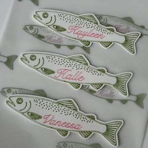 Könnte beinhalten: Drei gestanzte Aufkleber in Form von Forellen mit olivgrünen Umrissen und Details. Jeder Fisch trägt einen in rosa Schreibschrift geschriebenen Namen: Kayleen, Halle und Vanessa. Die Aufkleber sind auf einer transparenten Trägerfolie angeordnet.