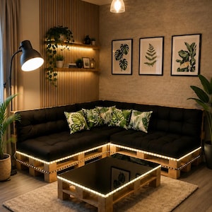 May include: An L-shaped sectional sofa made from light-coloured wooden pallets with black tufted cushions and green tropical leaf print throw pillows. A matching low pallet coffee table with a black glass top sits on a beige area rug.
