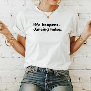 Puede incluir: Camiseta blanca con texto negro que dice "life happens. dancing helps."