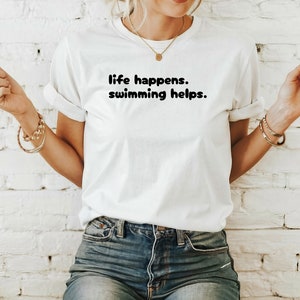 Puede incluir: Camiseta blanca con el texto "life happens. swimming helps."