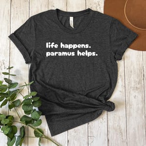 以下が含まれることがあります： 白い文字で「life happens. paramus helps.」とプリントされた濃いグレーのTシャツです。