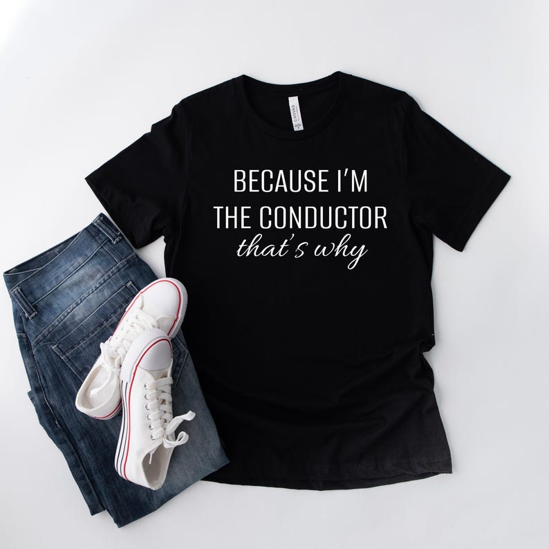 May include: Black t-shirt with white text that reads "Because I'm the conductor that's why". The t-shirt is lying flat on a white surface with a pair of blue jeans and white trainers beside it.
