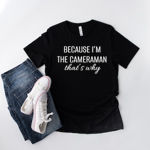 May include: Black crew neck t-shirt with the text "BECAUSE I'M THE CAMERAMAN that's why" in white. The shirt is paired with blue jeans and white trainers with red stripes.