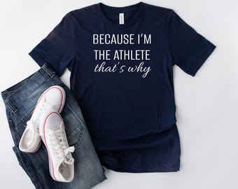 May include: A navy blue t-shirt with white text that reads "Because I'm the athlete that's why". The shirt is laid flat on a white surface with a pair of white sneakers and blue jeans.