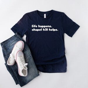 May include: A navy blue t-shirt with the text "life happens. chapel hill helps." White trainers with red detailing and blue jeans are next to the shirt. The shirt is made of a soft material.