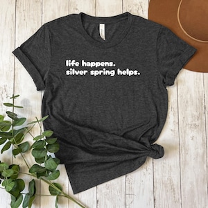 Peut inclure: Un t-shirt gris foncé avec le texte "life happens. silver spring helps." imprimé en blanc.