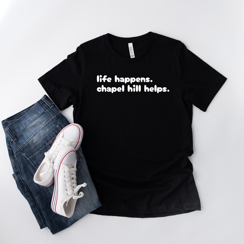May include: Black t-shirt with the text "life happens. chapel hill helps." in white. The shirt is paired with blue jeans and white trainers with red accents.