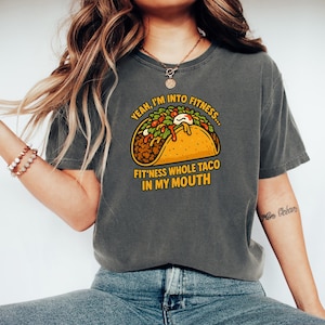 May include: Dark gray t-shirt featuring a colorful cartoon taco graphic. The taco is filled with various ingredients and the text reads "Yeah, I'm into fitness... Fit'ness whole taco in my mouth."