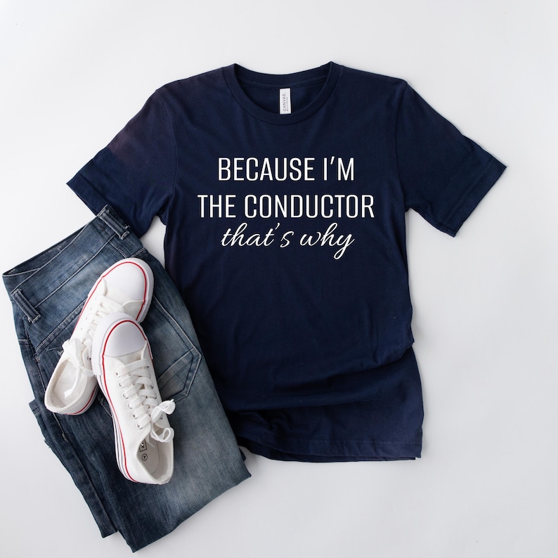 May include: A navy blue t-shirt with white text that reads "Because I'm the conductor that's why". The shirt is laid flat on a white surface with a pair of white trainers and blue jeans.