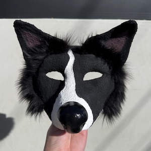 May include: A black and white animal mask featuring a textured surface, pointed ears with black fur trim, almond-shaped eye openings, and a prominent glossy black nose. The mask has a distinct white stripe running from the forehead to the nose.