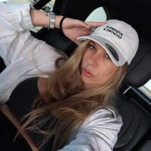 White baseball cap with the text "EXPENSIVE & DIFFICULT" in black. The cap has a gray brim. The person is wearing a black top and a white button-down shirt.