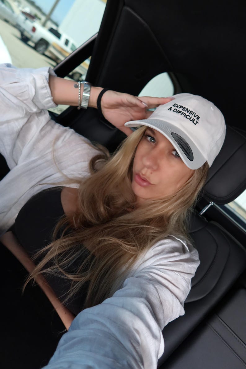 White baseball cap with the text "EXPENSIVE & DIFFICULT" in black. The cap has a gray brim. The person is wearing a black top and a white button-down shirt.