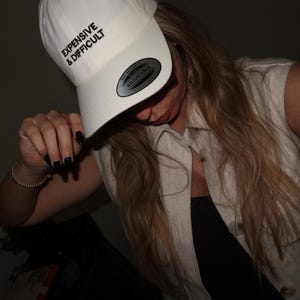 A white baseball cap with the text "EXPENSIVE & DIFFICULT" in black. The cap has a curved brim and an adjustable strap. The person in the image is wearing a sleeveless, light-colored shirt.