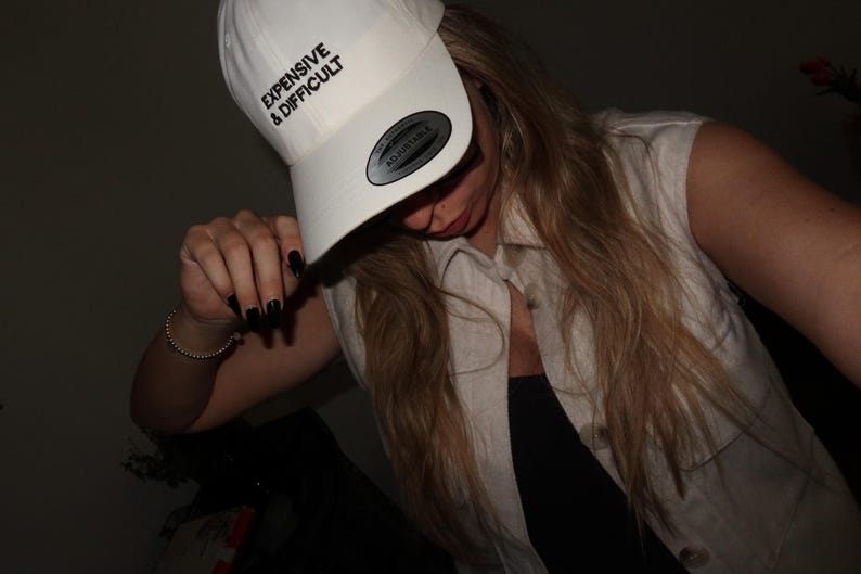 A white baseball cap with the text "EXPENSIVE & DIFFICULT" in black. The cap has a curved brim and an adjustable strap. The person in the image is wearing a sleeveless, light-colored shirt.
