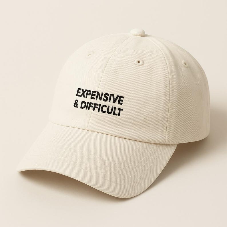 A cream-colored baseball cap with the text "EXPENSIVE & DIFFICULT" embroidered in black.