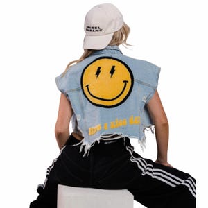 May include: Light blue denim vest with a large yellow smiley face patch and lightning bolts. The vest has a distressed hem and the words "Have a nice day" in yellow. A white baseball cap with black text is also visible.