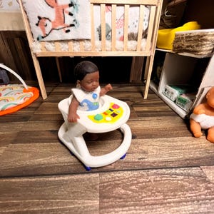 May include: A miniature doll in a white and blue bib sits in a white baby walker with colorful buttons. A wooden crib with a fox pillow and patterned bedding is in the background. The scene is set in a dollhouse nursery.