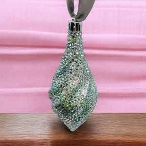 May include: A teardrop-shaped Christmas ornament covered in iridescent rhinestones. The ornament is silver with green and blue accents, and is suspended by a gray ribbon. The background is a pink fabric.