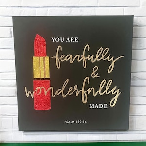 Può includere: Una tela nera con lettere di glitter dorate che recitano "You are fearfully & wonderfully made" con un'illustrazione di rossetto rosso. Il testo "Psalm 139:14" è in fondo alla tela.